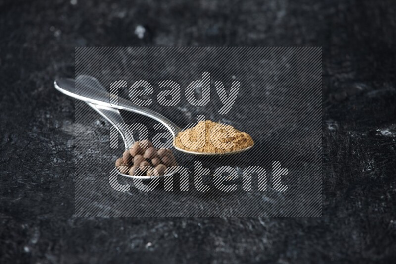 2 metal spoons full of allspice powder and whole balls on a textured black flooring