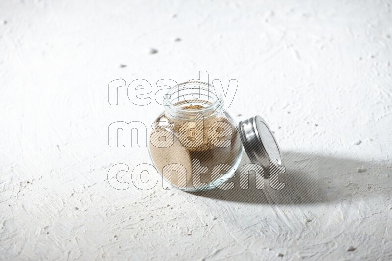 A glass spice jar full of cumin powder on textured white flooring