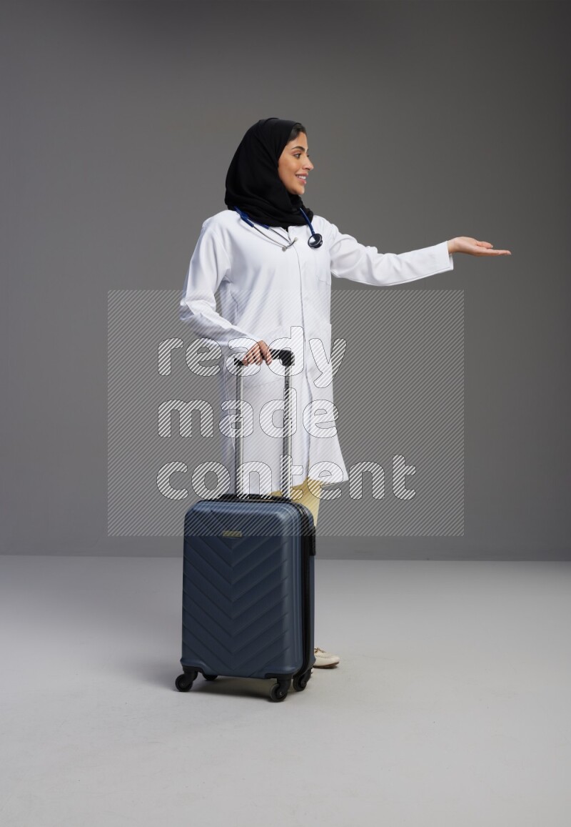 Saudi woman wearing lab coat with stethoscope standing holding Travel bag on Gray background