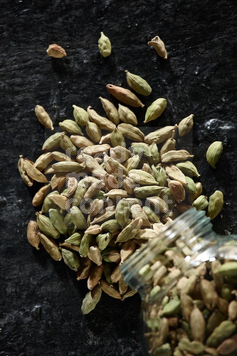 A flipped glass spice jar full of cardamom seeds on textured black flooring