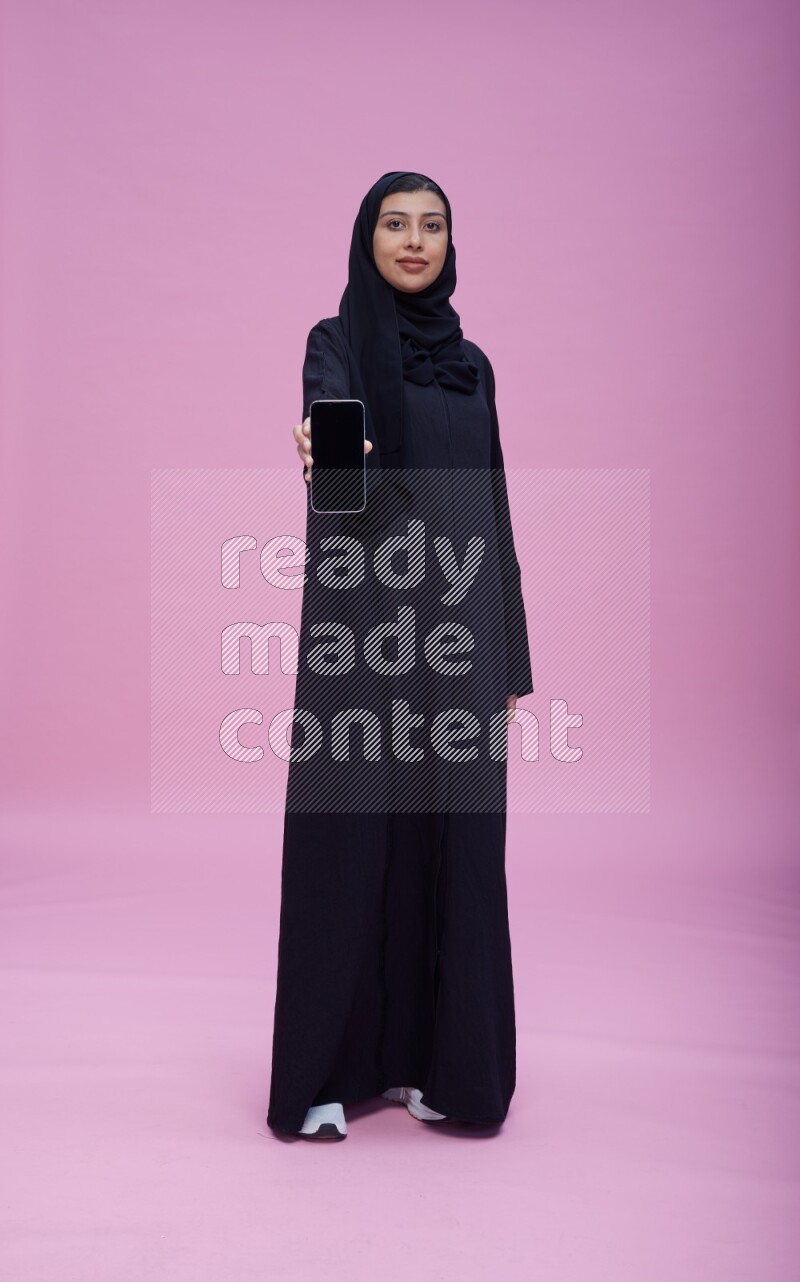 Saudi woman wearing Abaya standing showing phone to camera on pink background