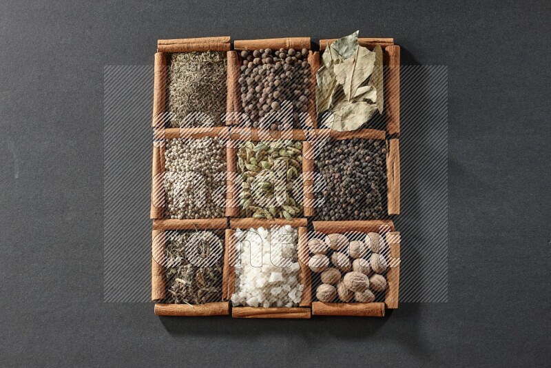 9 مربعات من أعواد القرفة ممتلئة بالهيل في المنتصف محاطة بجوزة الطيب والفلفل الأسود وورق الغار والبهارات والكمون والفلفل الأبيض والريحان المجفف والملح على أرضية سوداء