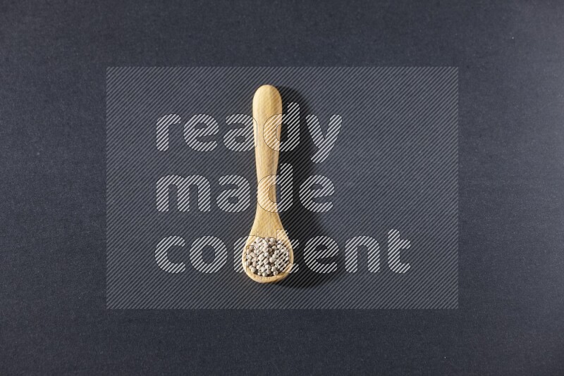 A wooden spoon full of white pepper beads on black flooring