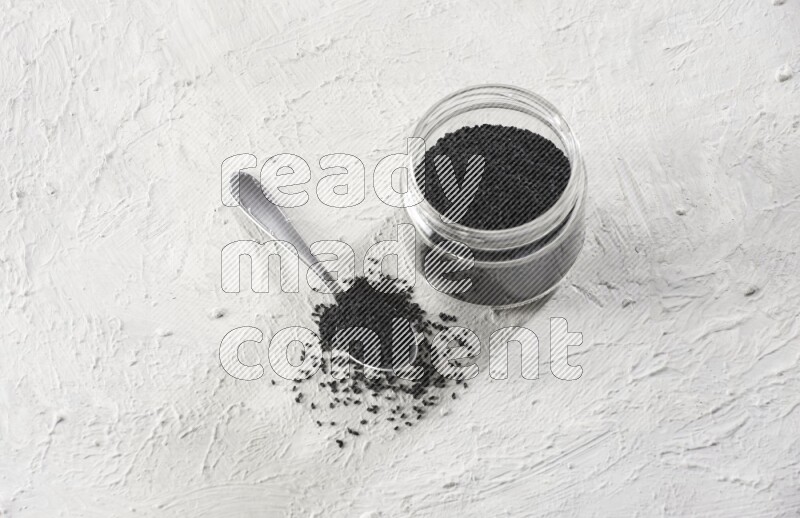 A glass jar and a metal spoon full of black seeds on a textured white flooring