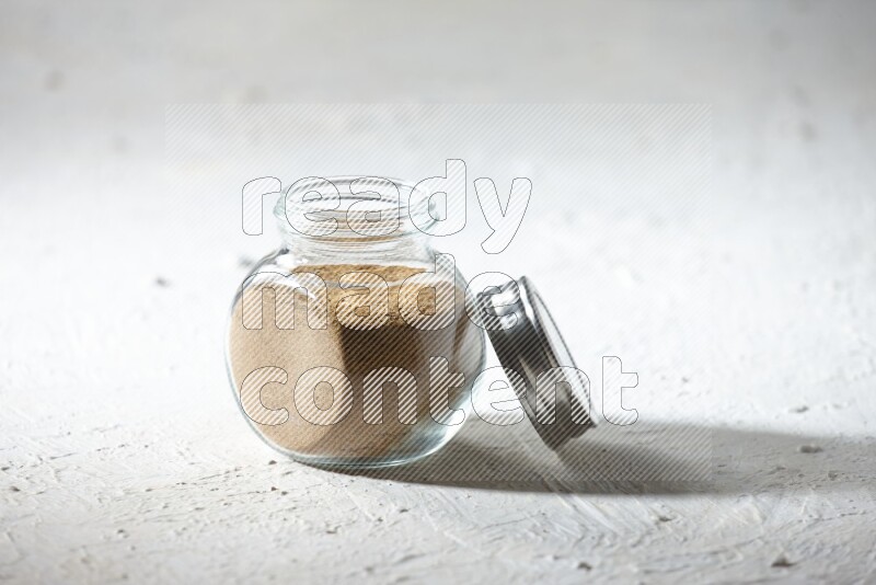 A glass spice jar full of cumin powder on textured white flooring