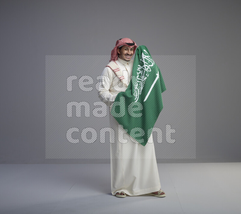 A saudi man standing wearing thob and red shomag holding big saudi flag on gray background