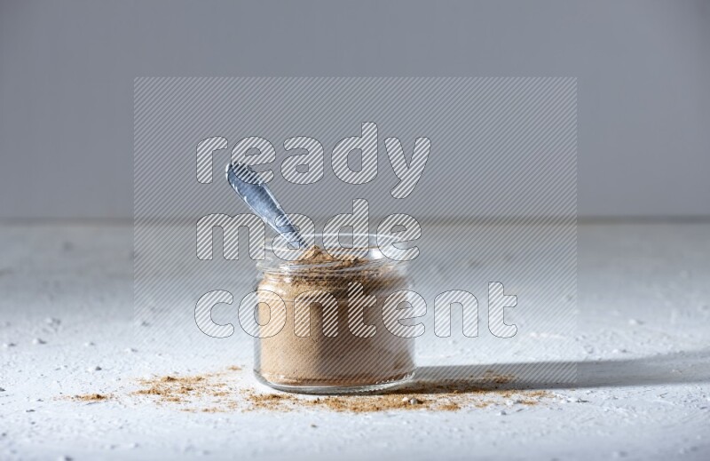 A glass jar and a metal spoon full of allspice powder on a textured white flooring