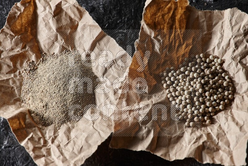 White pepper beads and powder in 2 crumpled paper on textured black flooring