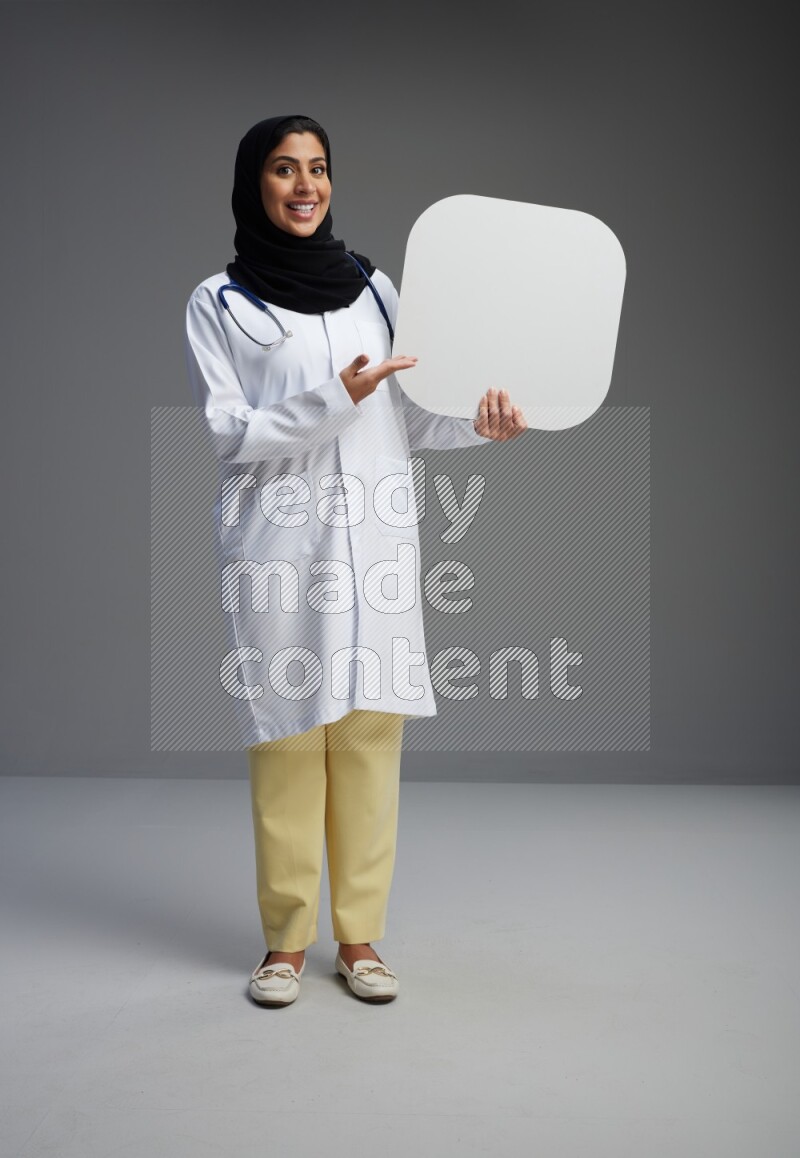 Saudi woman wearing lab coat with stethoscope standing holding social media sign on Gray background