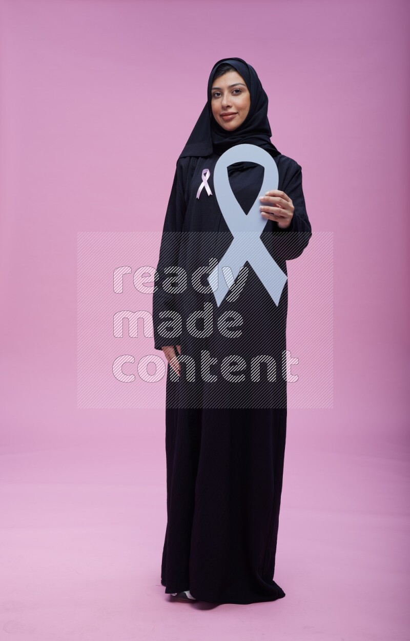 Saudi woman wearing pink ribbon on abaya standing holding awareness ribbon on plain pink background