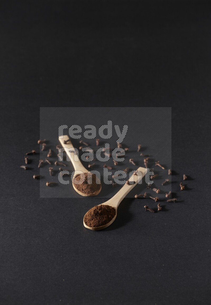 2 wooden spoons full of cloves powder with spreaded whole cloves on a black flooring