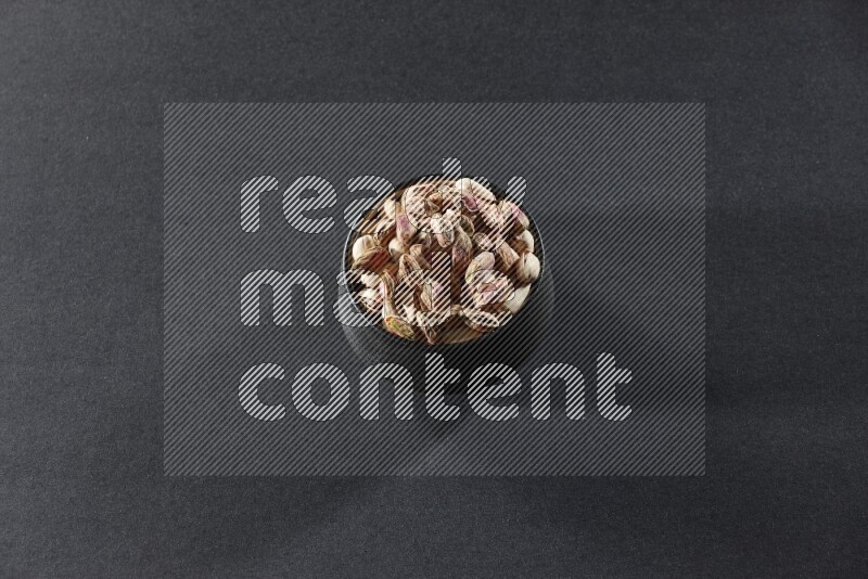 A black pottery bowl full of peeled pistachios on a black background in different angles