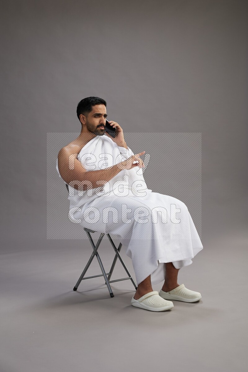 A man wearing Ehram sitting on chair talking on the phone on gray background