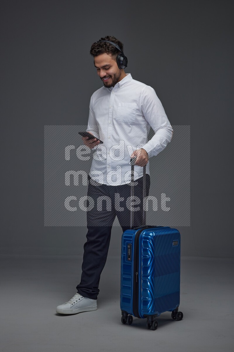 A man wearing smart casual using his phone while wearing headphone and holding luggage eye level on a gray background