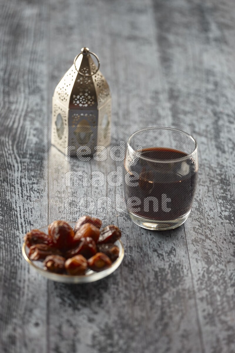A silver lantern with different drinks, dates, nuts, prayer beads and quran on grey wooden background