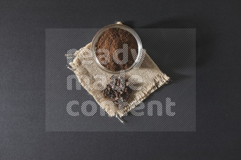 A glass bowl full of cloves powder with cloves grains on a burlap piece on a black flooring