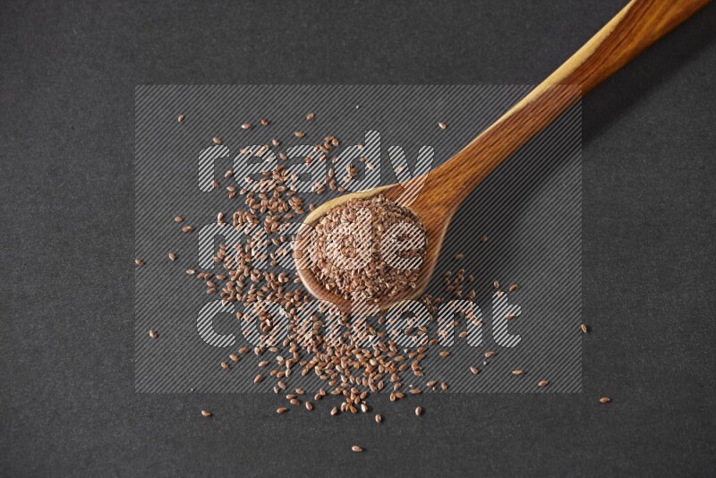 A wooden ladle full of flaxseeds and seeds spread beside it on a black flooring