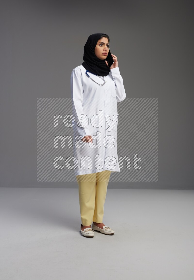Saudi woman wearing lab coat with stethoscope standing talking on phone on Gray background