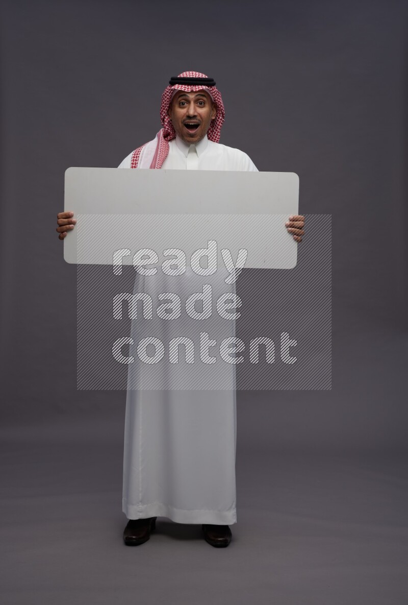 Saudi man wearing thob and shomag standing holding board on gray background