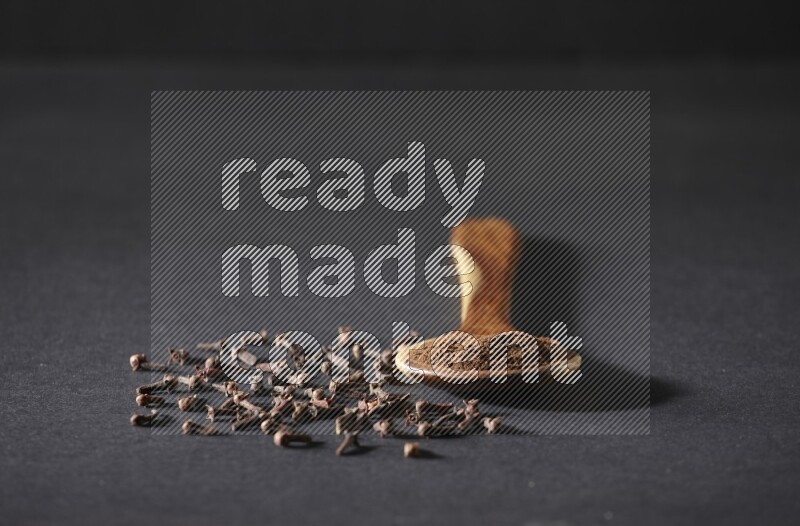 A wooden ladle full of cloves powder and some of whole cloves around it on a black flooring