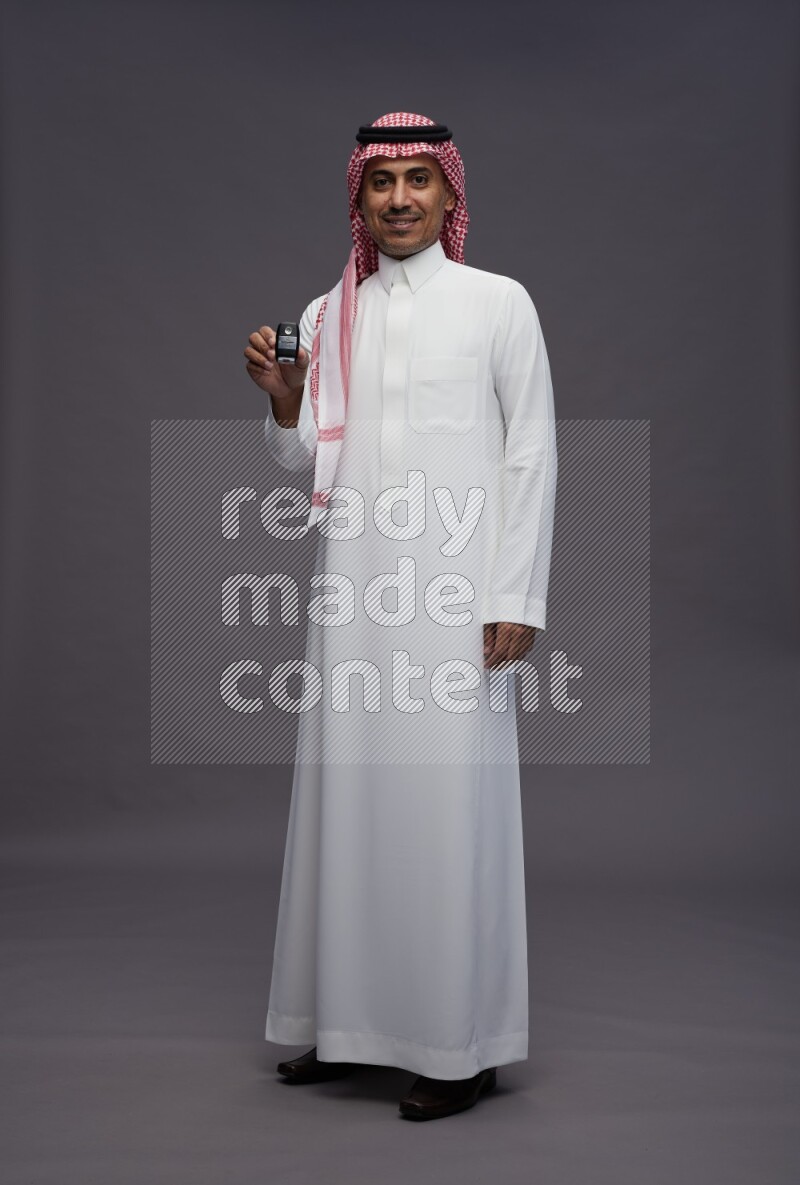 Saudi man wearing thob and shomag standing holding key car on gray background