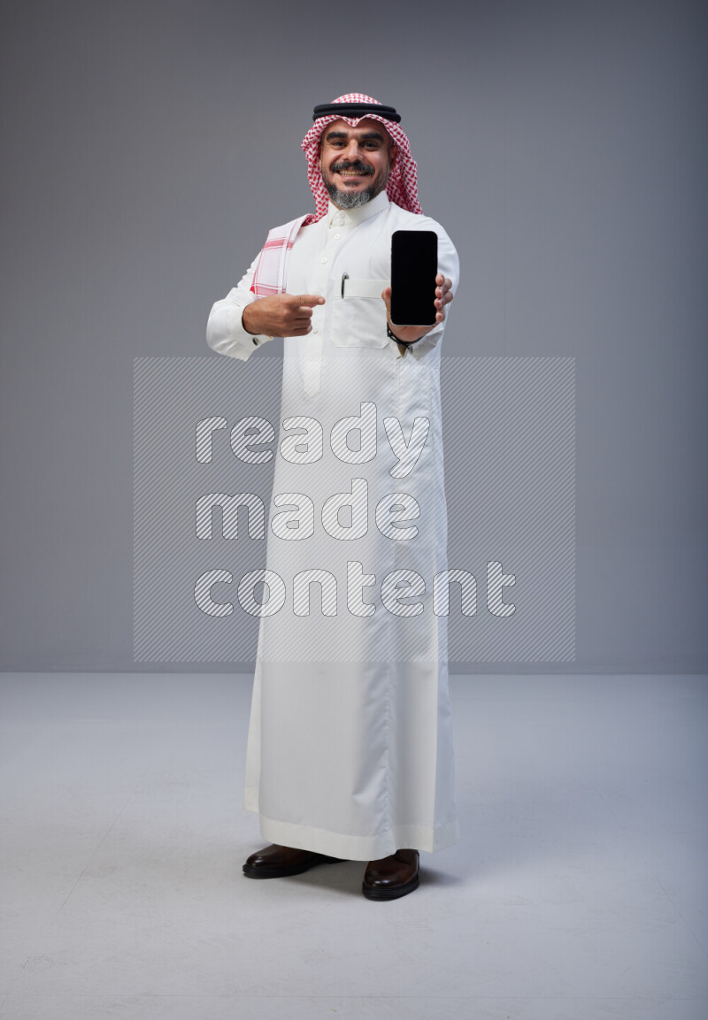 Saudi man Wearing Thob and red Shomag standing showing phone to camera on Gray background