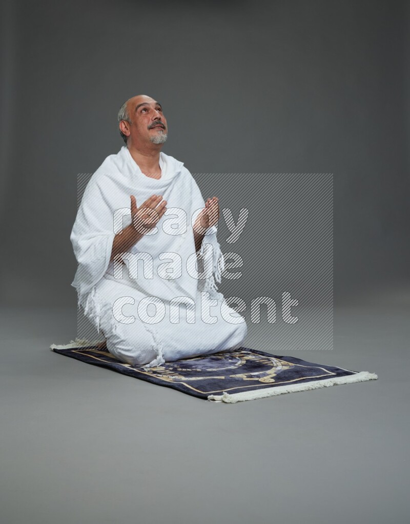 A man wearing Ehram sitting on prayer mat dua'a on gray background