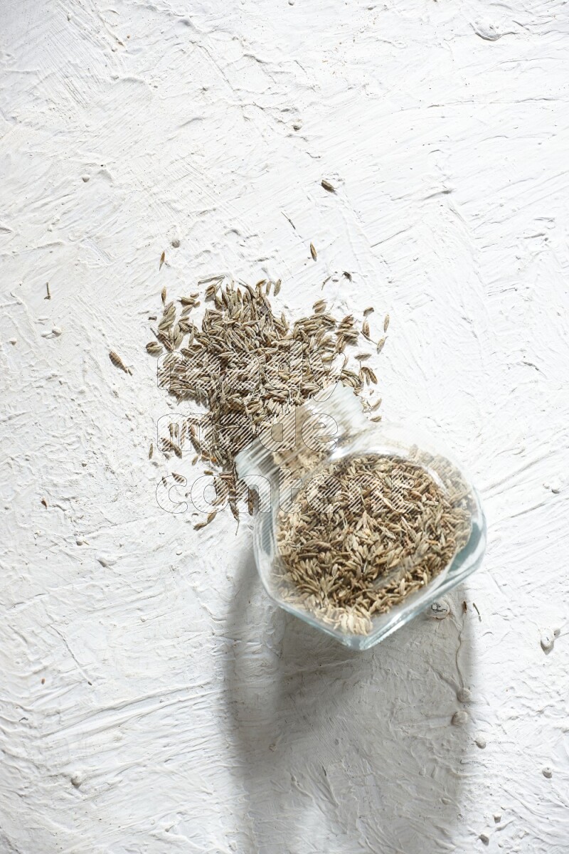 A flipped glass spice jar full of cumin seeds and the seeds spilled out on textured white flooring