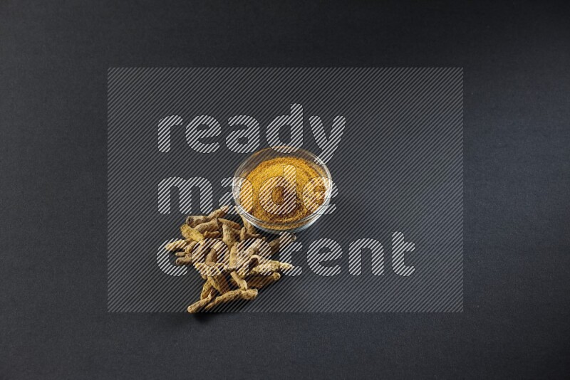 وعاء زجاجي ممتلئ ببودرة الكركم وأصابع الكركم المجففة بجانبه على خلفية سوداء