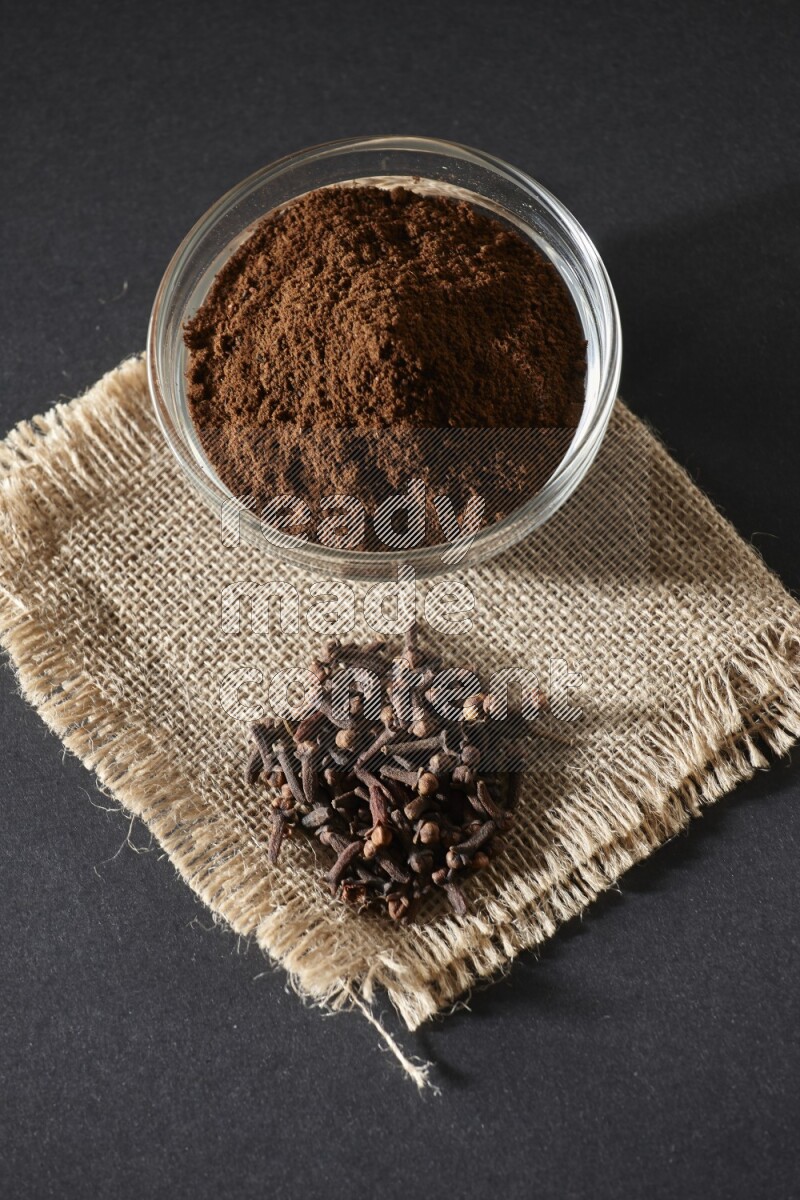 A glass bowl full of cloves powder with cloves grains on a burlap piece on a black flooring