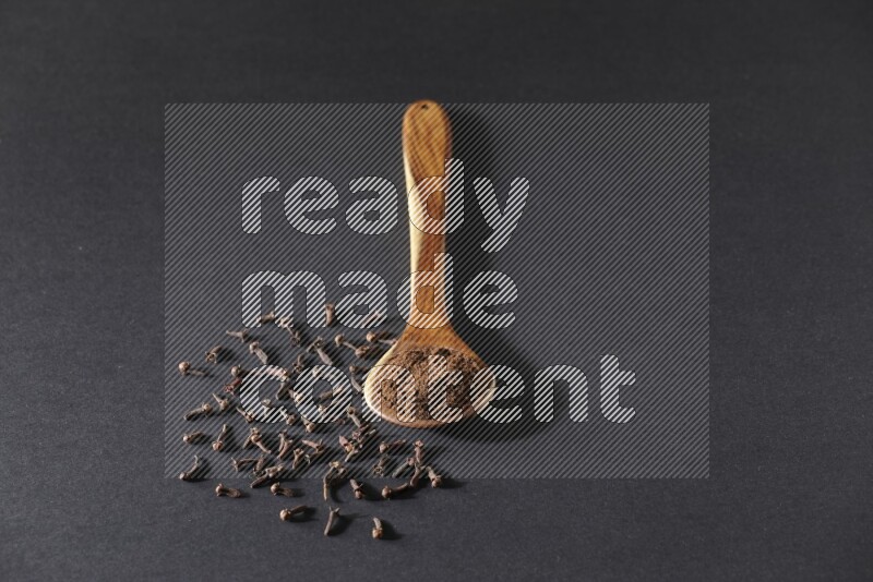 A wooden ladle full of cloves powder and some of whole cloves around it on a black flooring