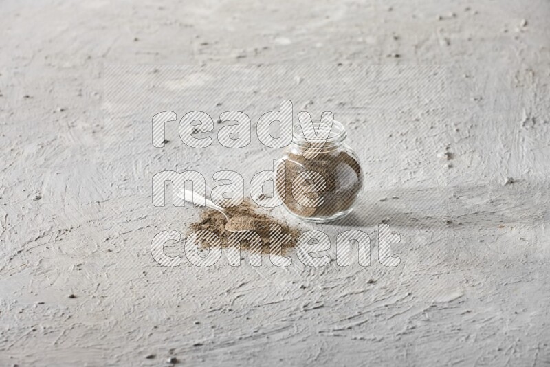 A glass spice jar full of black pepper powder and metal spoon full of it on textured white flooring