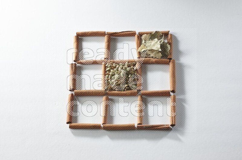 9 squares of cinnamon sticks full of cardamom in the middle surrounded by nutmeg, cinnamon, bay laurel leaves, cloves, cumin, dried ginger, dried basil and star anise on white flooring