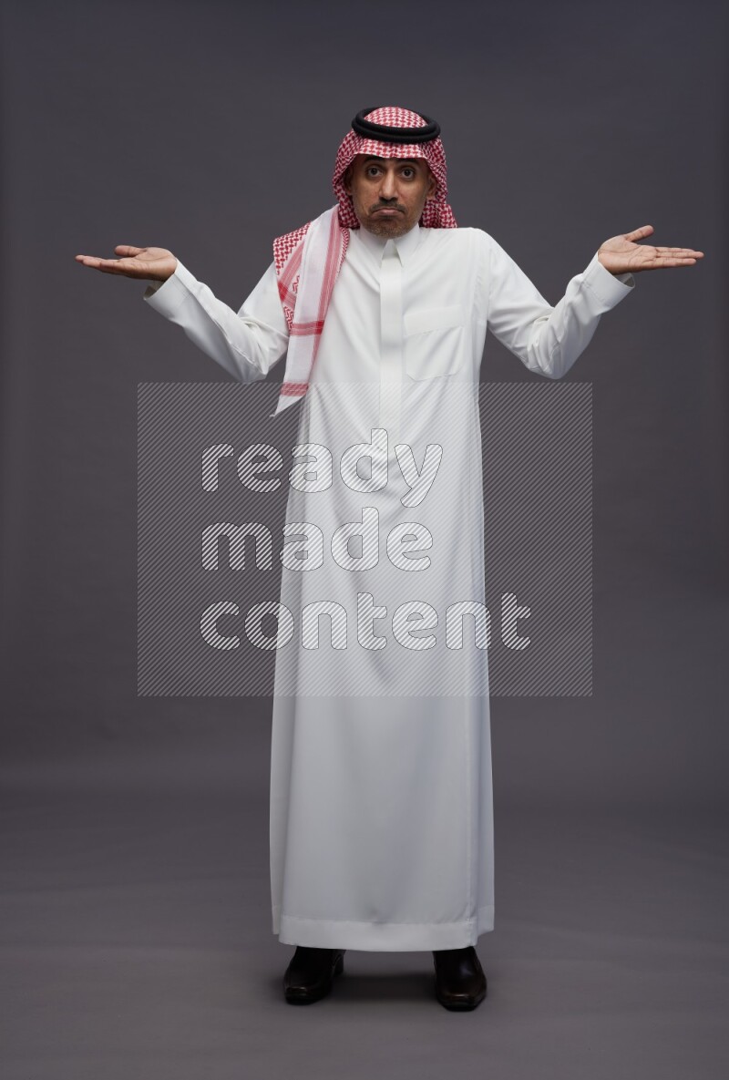 Saudi man wearing thob and shomag standing interacting with the camera on gray background