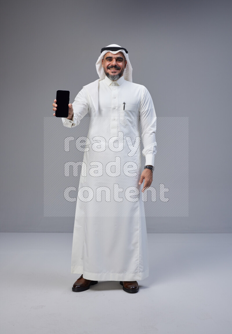 Saudi man Wearing Thob and white Shomag standing showing phone to camera on Gray background