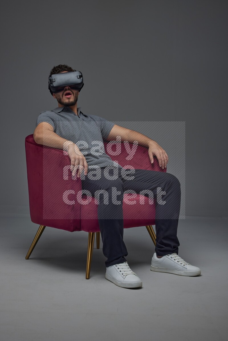 A man wearing casual and wearing VR while sitting on a burgundy chair eye level on a gray background