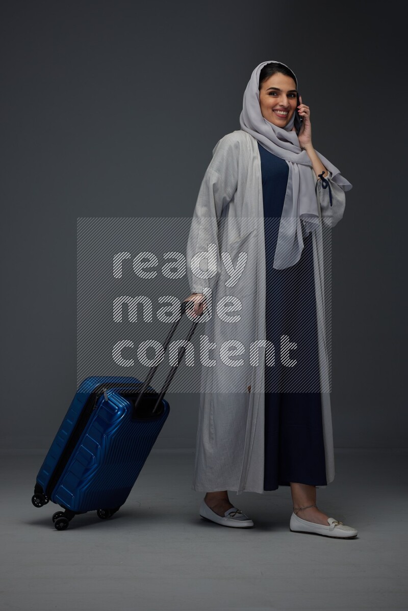 A Saudi woman wearing a light gray Abaya and head scarf standing and talking in the phone while holding shopping bags eye level on a grey background