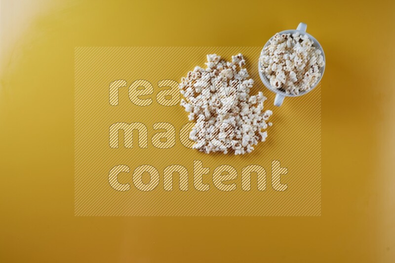 A white ceramic bowl full of popcorn with popcorn beside it on a yellow background in a top view shot