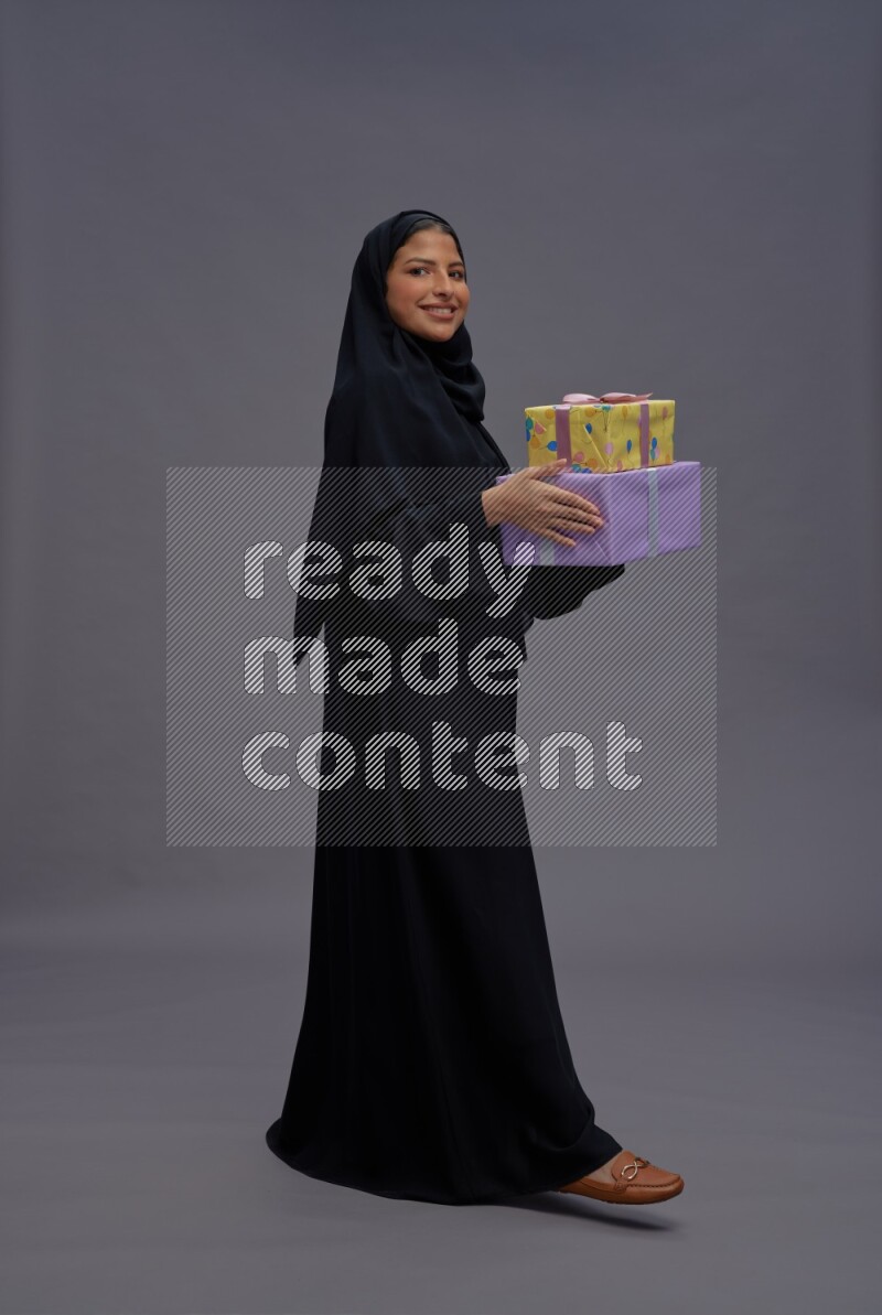 Saudi woman wearing Abaya standing holding gift box on gray background