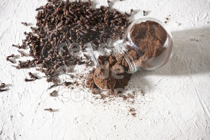 A flipped glass spice jar and a metal spoon full of cloves powder and powder came out of the jar with cloves spread on textured white flooring
