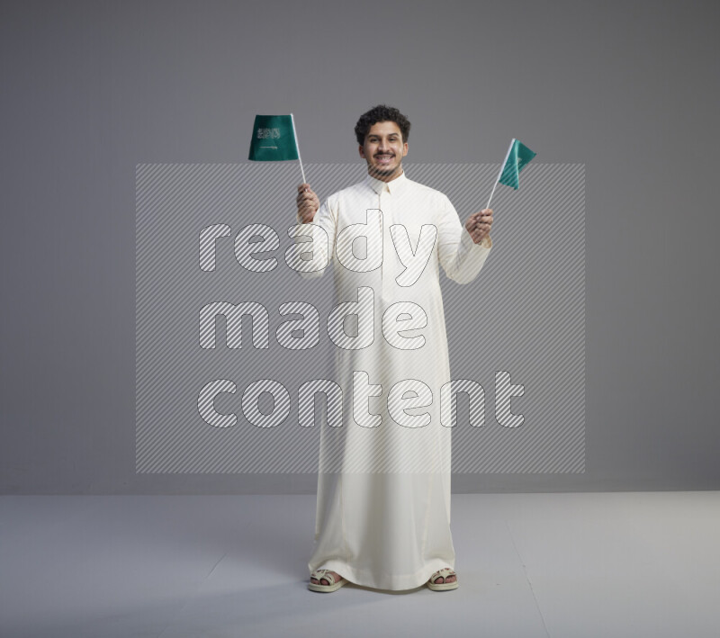 A Saudi man standing wearing thob holding small Saudi flag on gray background