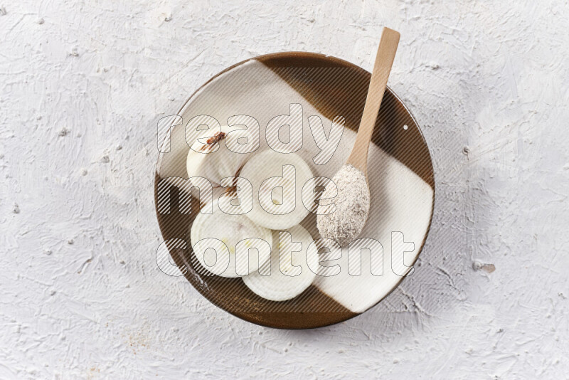 A wooden spoon full of onion powder with sliced onions, all on a pottery plate on white background