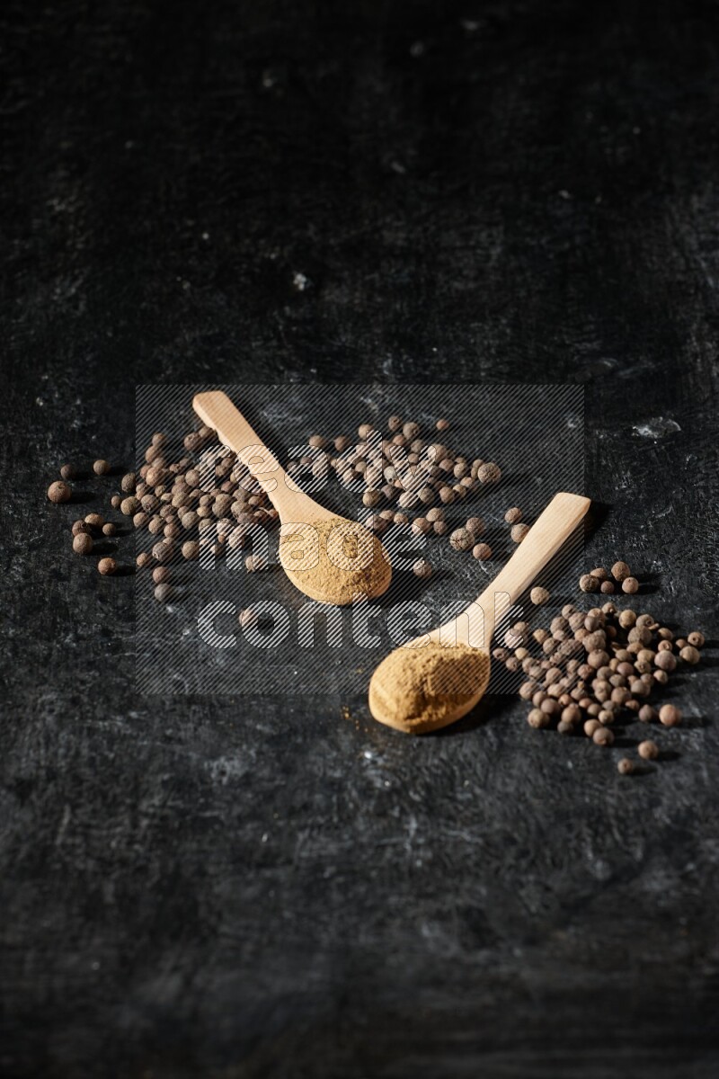 Wooden spoons full of all spice powder and allspice whole balls beside it on a textured black flooring