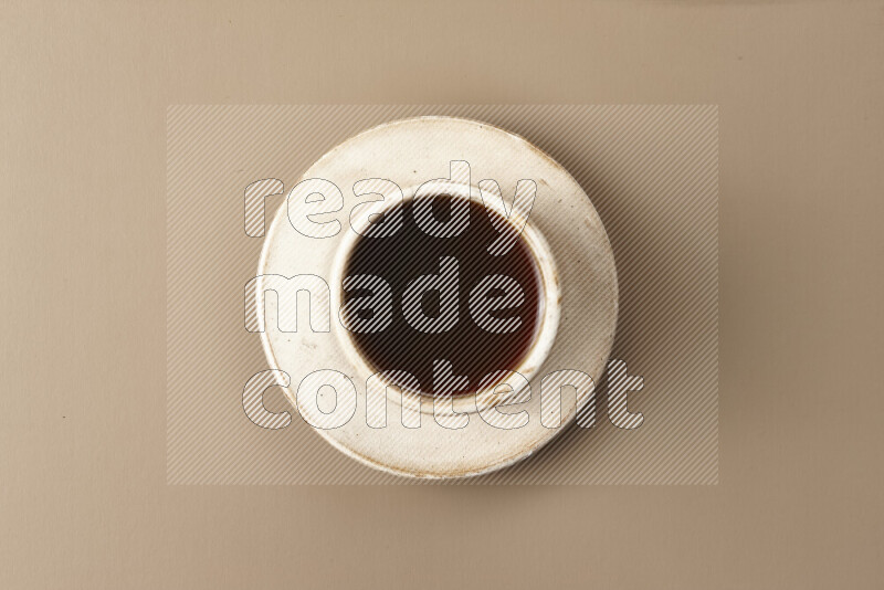 A beige pottery cup of coffee surrounded by roasted coffee beans on beige background