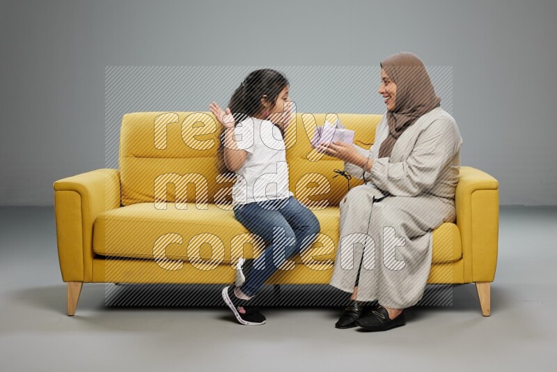 A girl sitting on a yellow sofa and giving a gift to her mother on gray background