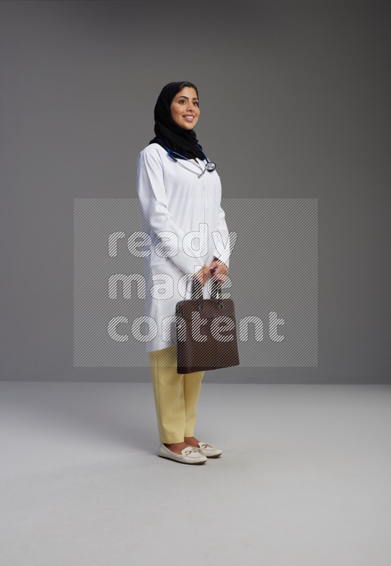 Saudi woman wearing lab coat with stethoscope standing holding bag on Gray background