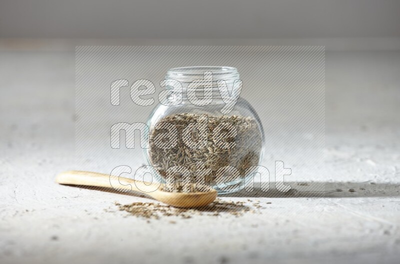 A glass spice jar and wooden spoon full of cumin seeds on textured white flooring