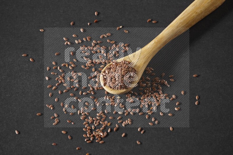 A wooden spoon full of flaxseeds and seeds spread beside it on a black flooring