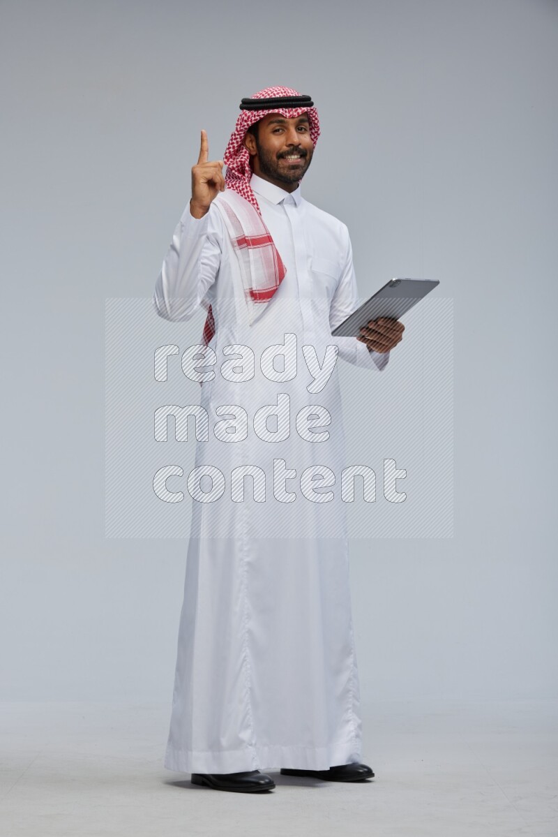 Saudi man Wearing Thob and shomag standing working on tablet on Gray background