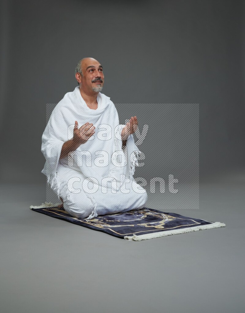 A man wearing Ehram sitting on prayer mat dua'a on gray background